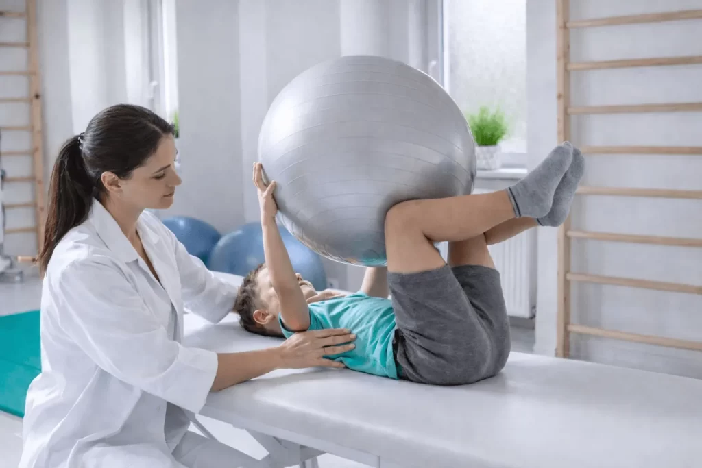 O nás - REHANEST dětská rehabilitace Praha Barrandov – fyzioterapeutka pomáhá chlapci při cvičení s velkým rehabilitačním míčem na lehátku dětské rehabilitační ambulanci.